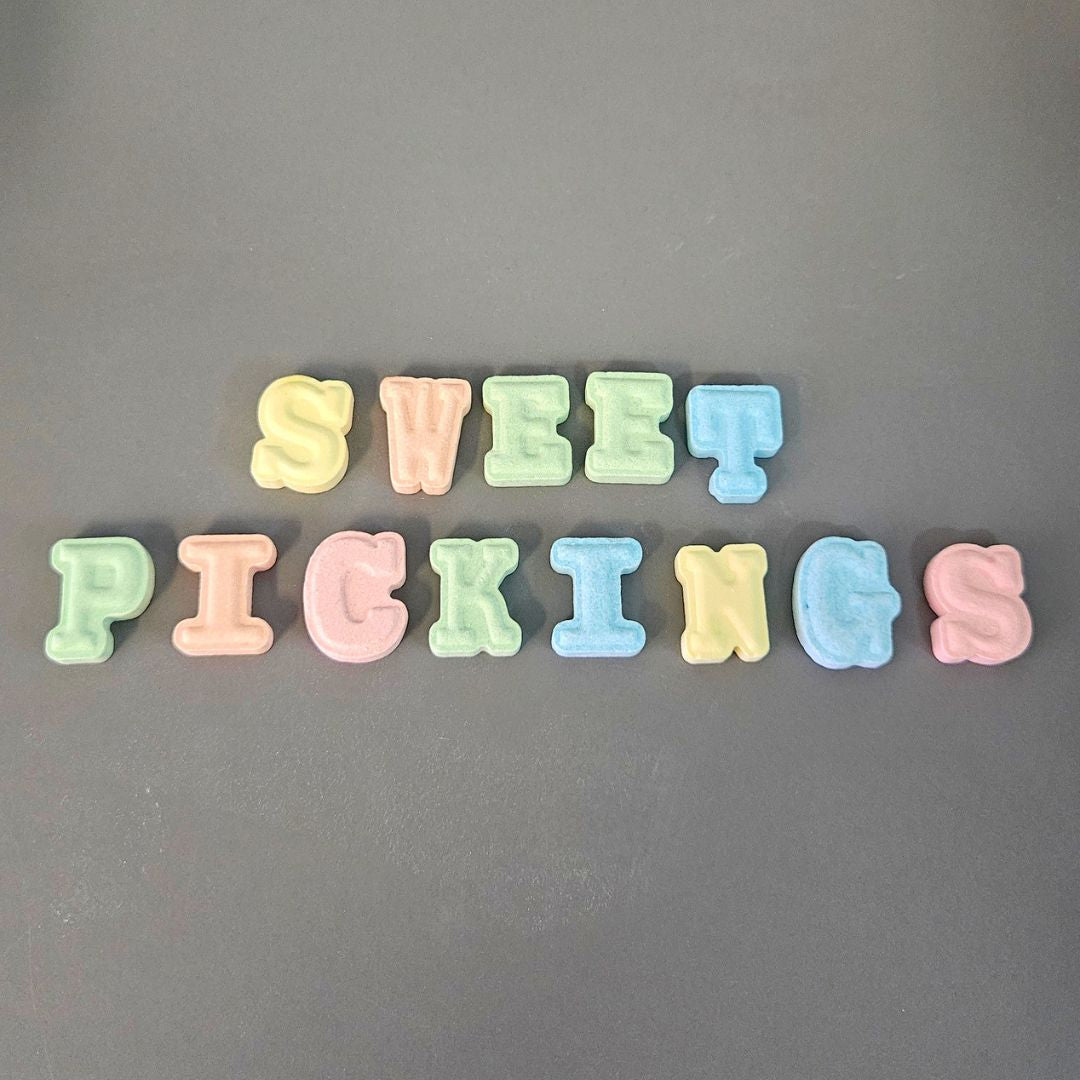 The word 'SWEET PICKINGS' spelled out in pastel-colored, 3D alphabet letters arranged in two lines on a dark gray background.