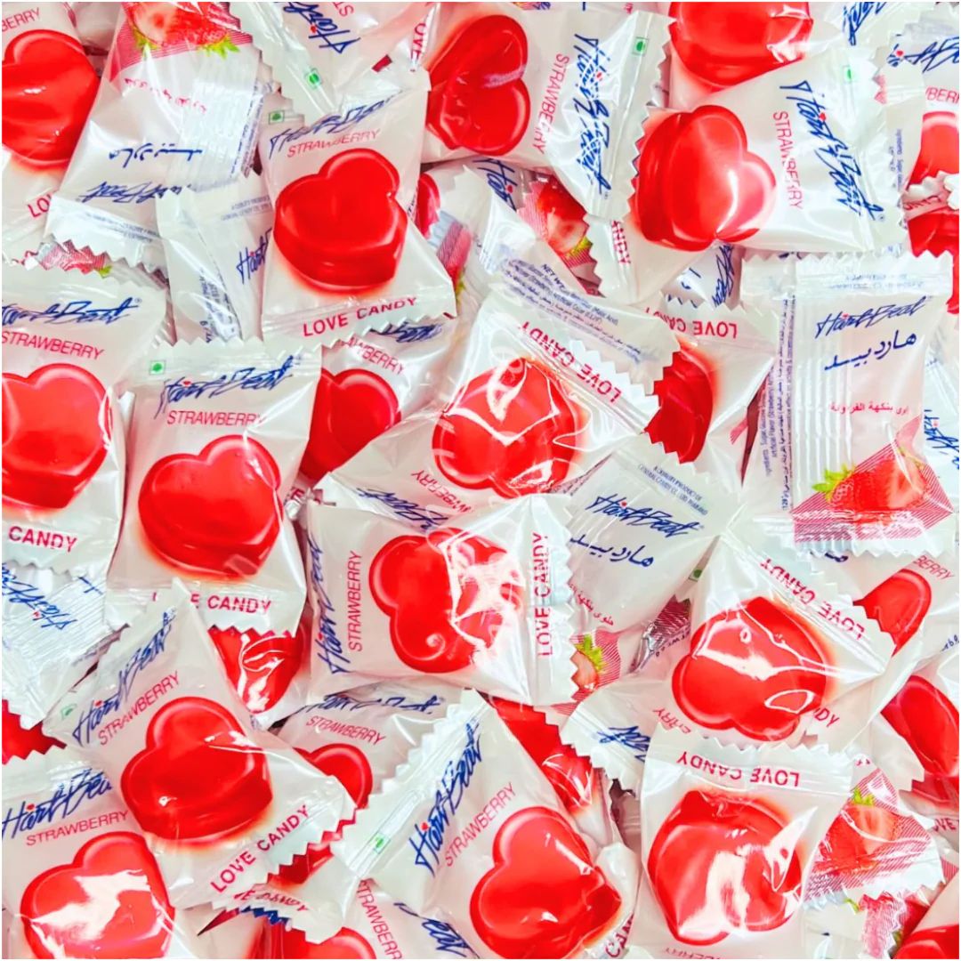 A pile of individually wrapped heart-shaped strawberry candy, showing the red candy pieces through clear plastic packaging with white wrappers and blue text.
